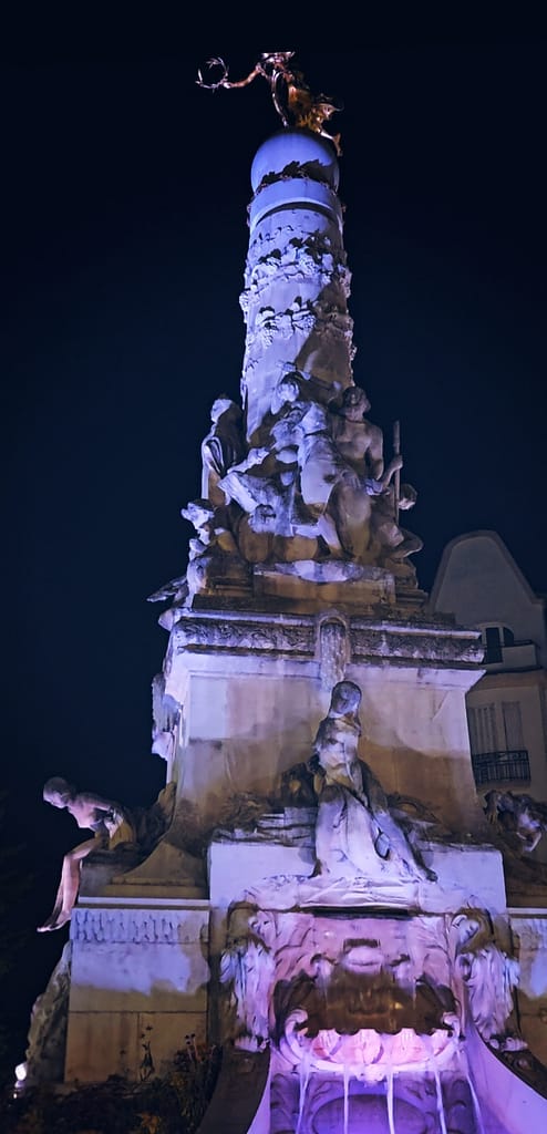 Fontaine Subé in Reims lit up at night, seen from below with statues and golden figure on top.