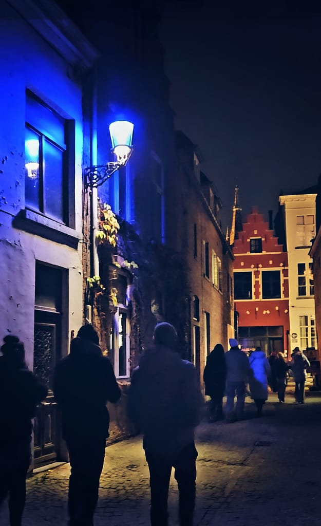 Calle estrecha en Brujas iluminada con luces azules del recorrido Winter Glow, con gente paseando de noche.