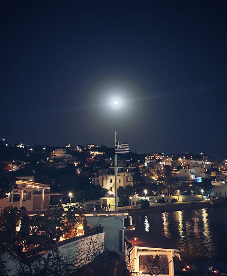 Night view of Chora, Mykonos with the Greek flag and town lights reflecting on the water