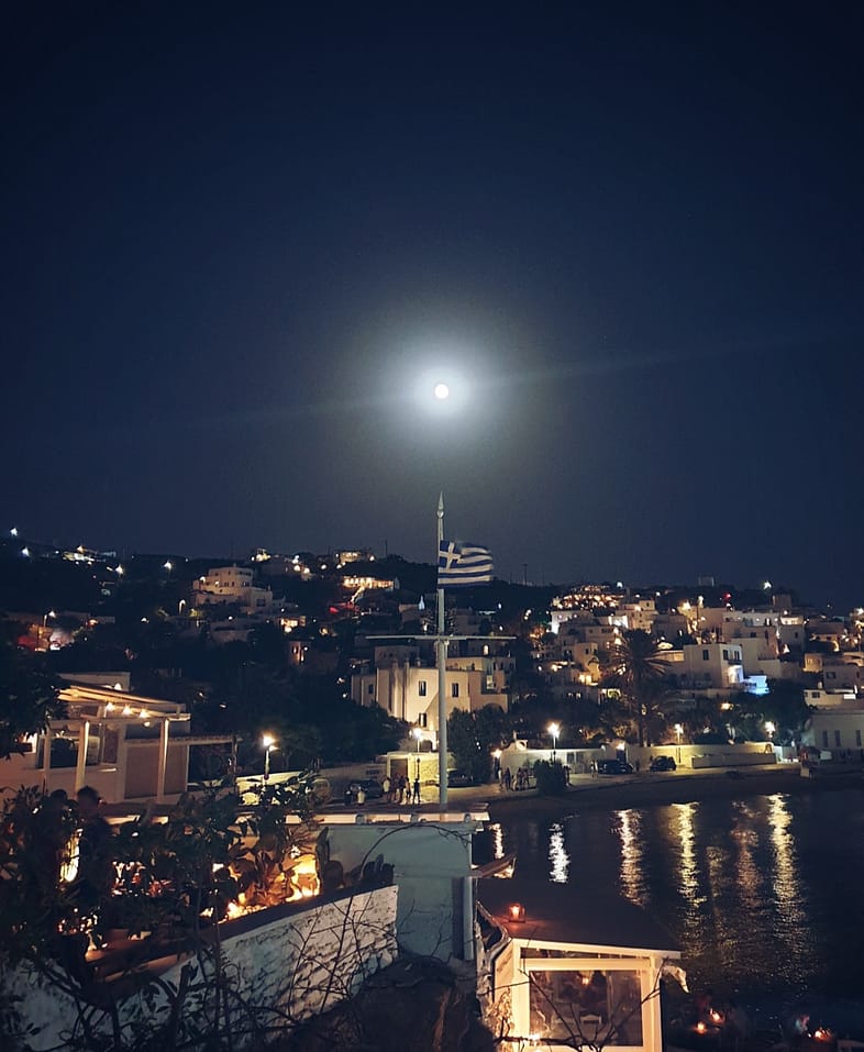 Vista nocturna de Chora Mykonos con bandera griega, luces del pueblo y reflejos en el agua